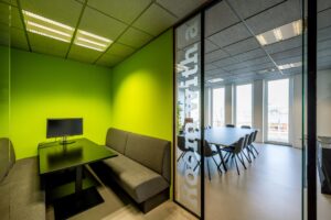 Moderne vergaderruimte aan de De Ruijterkade met felgroene wand, zithoek met monitor en glazen tussenwand richting een grotere vergaderzaal.
